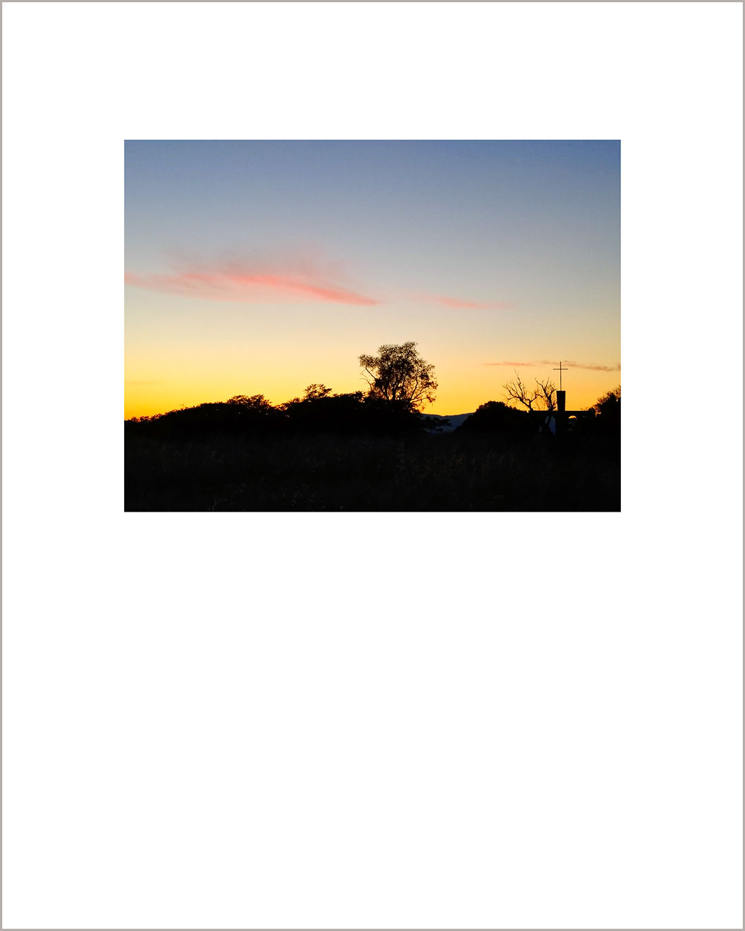 Fotografía horizontal de un atardecer, con el suelo negro y la silueta de un árbol y una cruz, el cielo hace un degradado del amarillo al azul grisáceo con dos nubes rosas en medio.