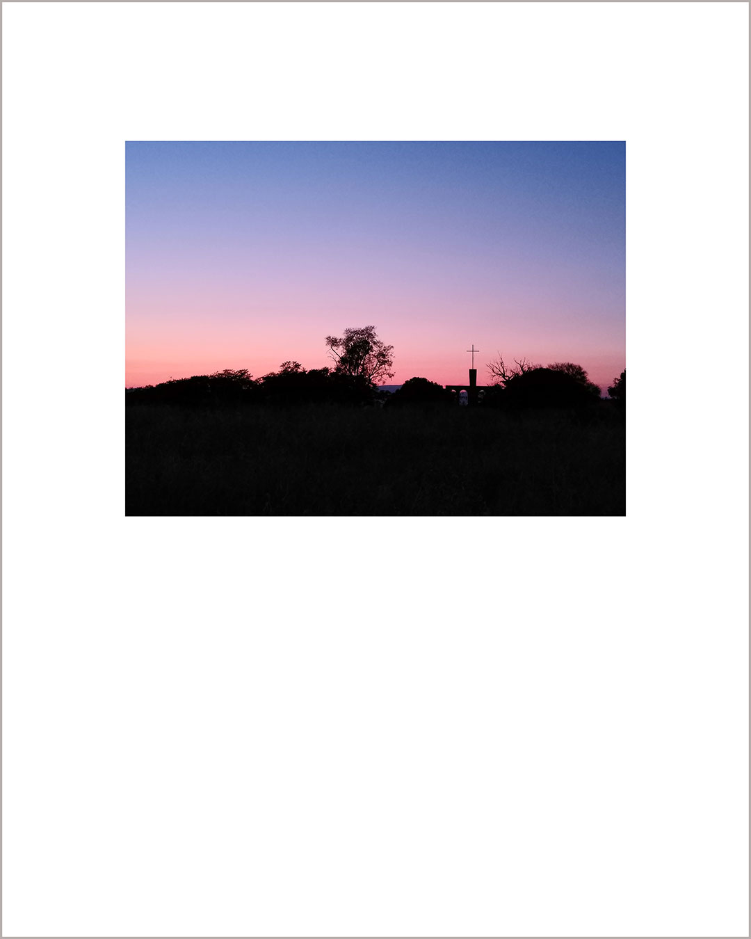 Fotografía horizontal de un atardecer, con el suelo negro y la silueta de un árbol y una cruz, el cielo hace un degradado del rosa al añil.
