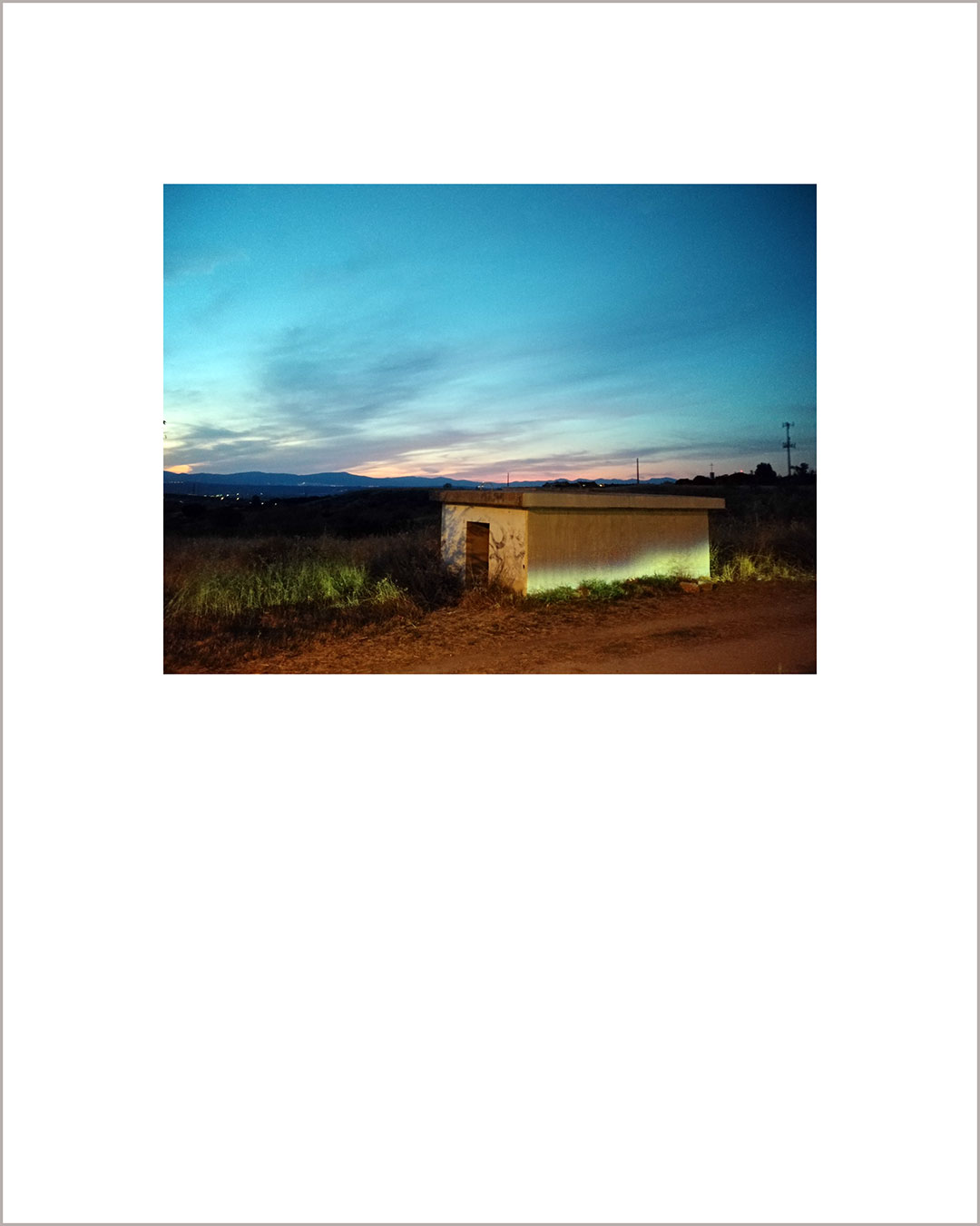 Fotografía horizontal de un atardecer, en primer término hay una caseta iluminada por las luces de un coche.