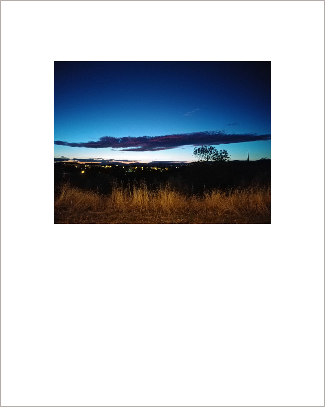 Fotografía horizontal de un atardecer, en el suelo se ve una franja de paja amarilla sobre un negro, hay luces en el horizonte, el cielo es añil y destaca una gran nube horizontal en azul oscuro.