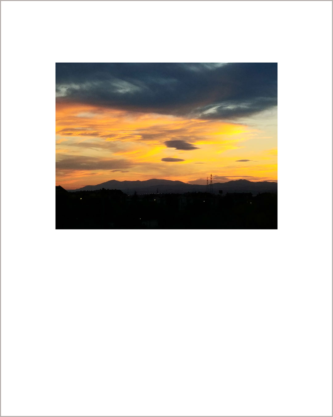 Fotografía horizontal de un atardecer, el suelo es negro y al fondo se ve la silueta de unas montañas, el cielo está cubierto de unas nubes naranjas y hay otras nubes gris oscuro en la parte superior.