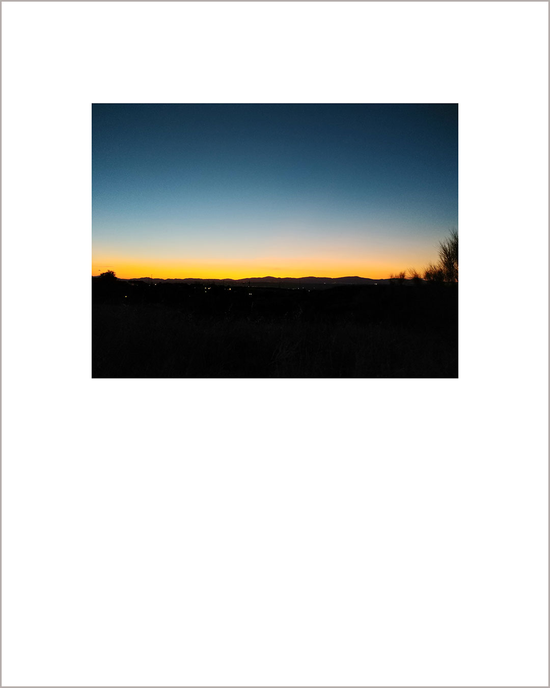Fotografía horizontal de un atardecer, el suelo es negro y en el horizonte se dibuja una línea naranja que degrada hacia el cielo azul plomizo.