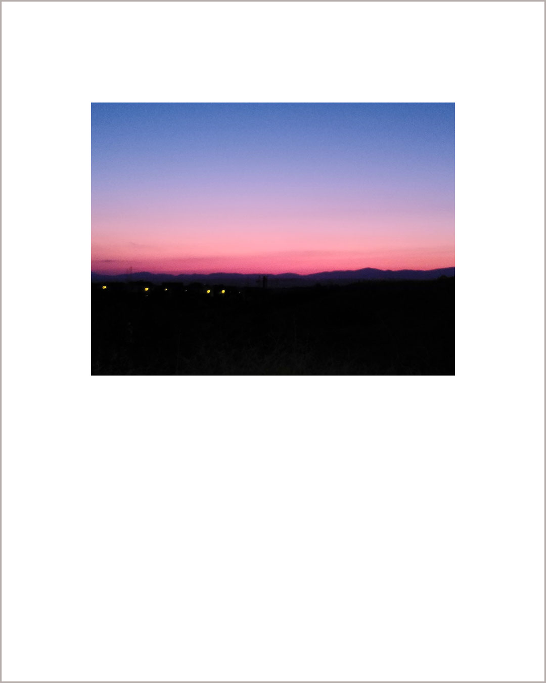 Fotografía horizontal de un atardecer, en el suelo negro destacan unas luces lejanas, el cielo hace un degradado del rosa al azul.