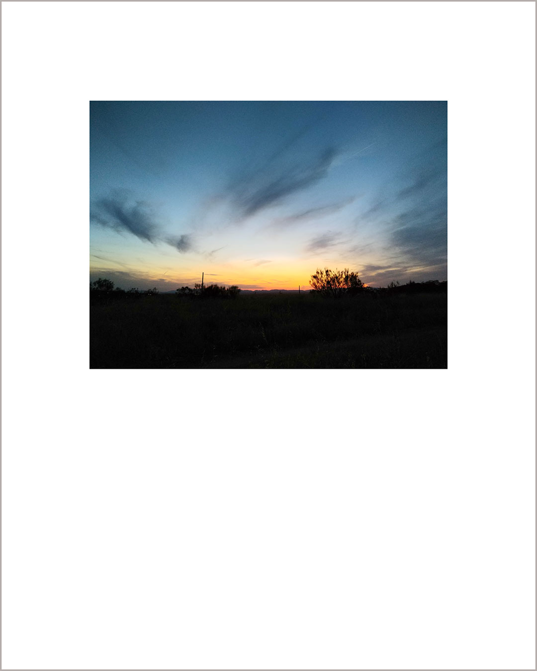 Fotografía horizontal de un atardecer, suelo negro y cielo azul con algunas nubes suaves en color plomizo.