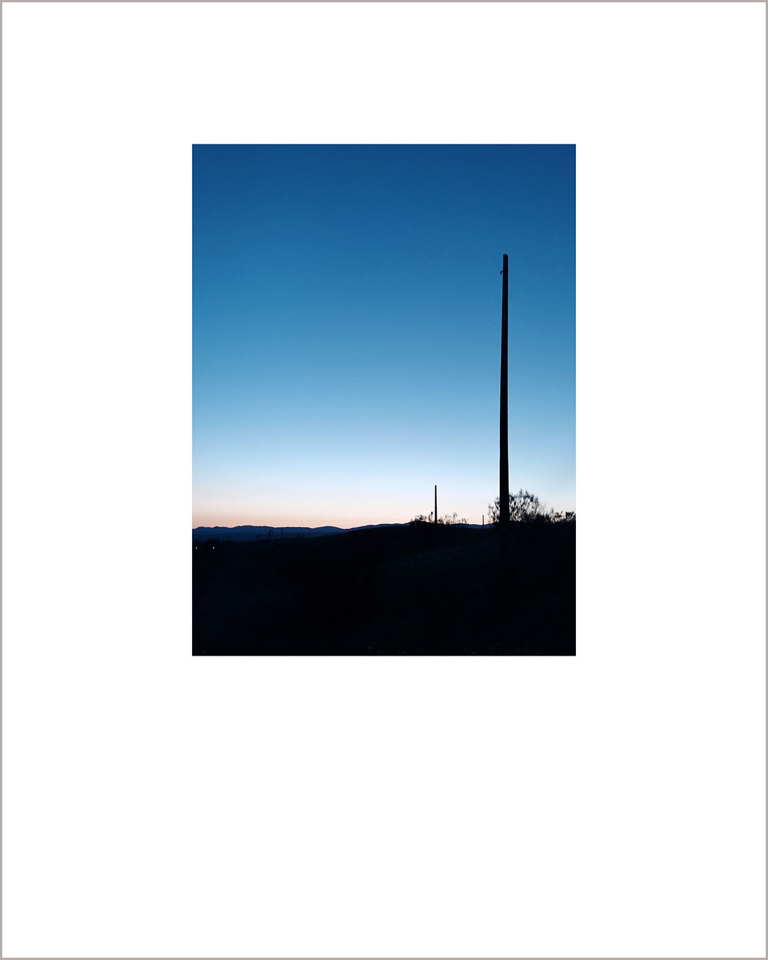 Fotografía vertical de un atardecer, con el suelo negro y la silueta de un poste a la derecha, el cielo es azul.
