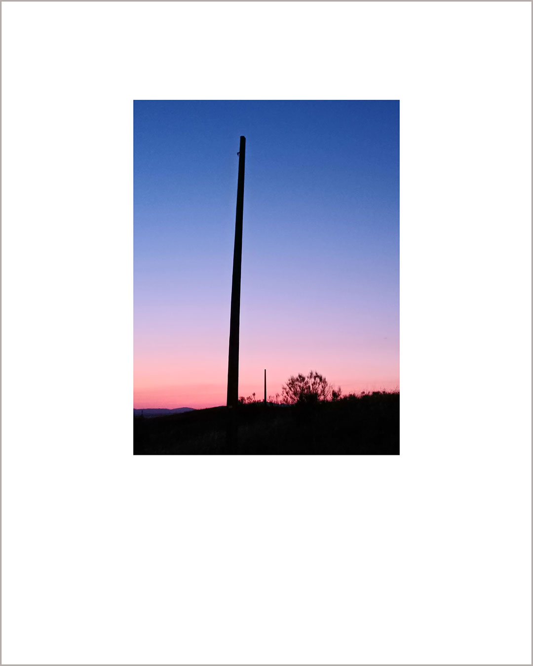 Fotografía vertical de un atardecer, el suelo es negro y hay un gran poste en el centro, el cielo está degradado del rosa al añil.