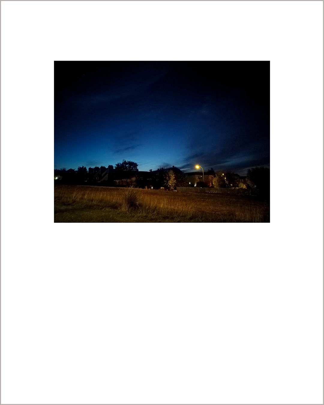 Fotografía horizontal de un anochecer, en el suelo se ve la hierba seca y una luz a lo lejos, el cielo es añil y negro.