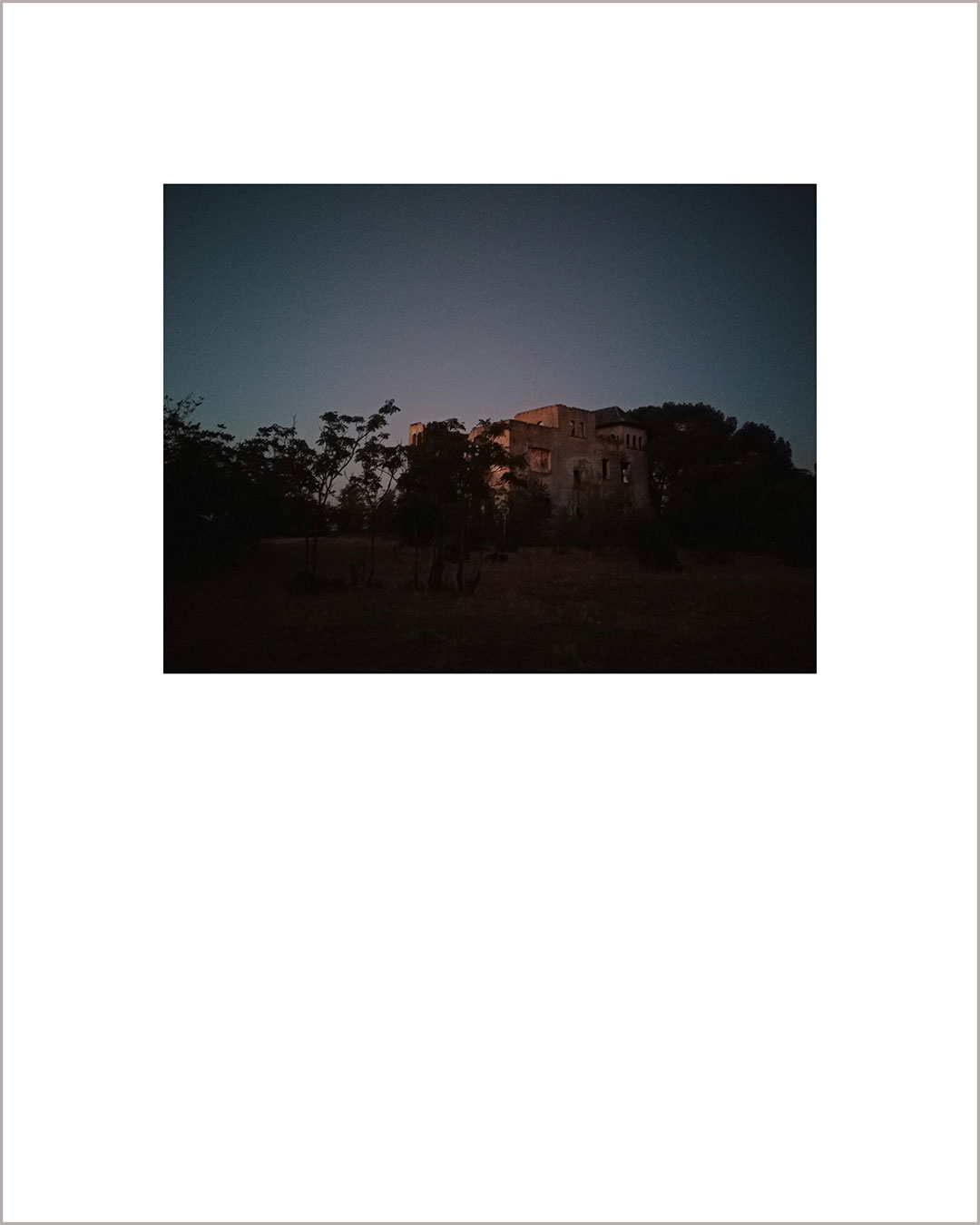 Fotografía horizontal de un anochecer, aparce las ruinas de una casa y unos árboles negros en primer plano, el cielo es color plomo.